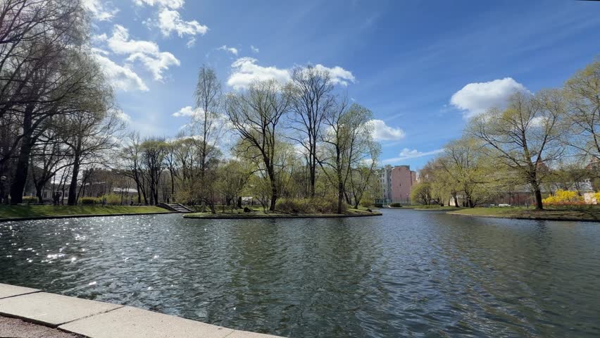 Tranquil park scene featuring a pond, lush trees, and buildings under a clear blue sky with fluffy white clouds reflecting on the water