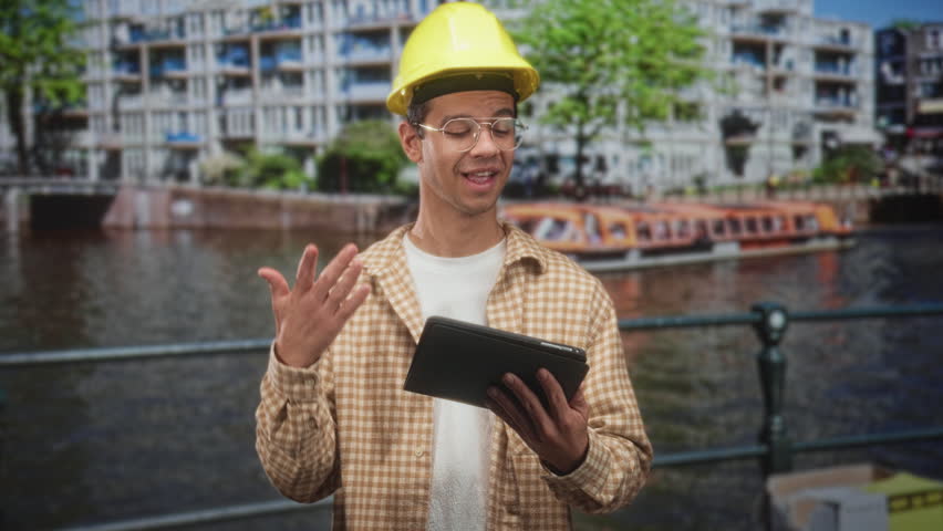 Man in yellow hardhat holds tablet and gestures with hand by riverside building as architect on site; planning focus.