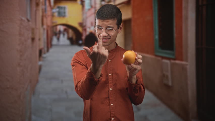 Man holding an orange while showing middle finger on a narrow cobbled street in an urban alley, wearing glasses and orange shirt; playful defiance.