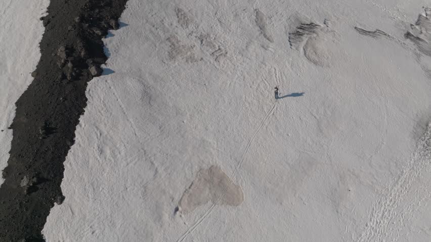Aerial view of a lone hiker walking across a vast snowfield next to a dark volcanic ridge near Rifugio Sapienza in Nicolosi, Sicilia, Italy.