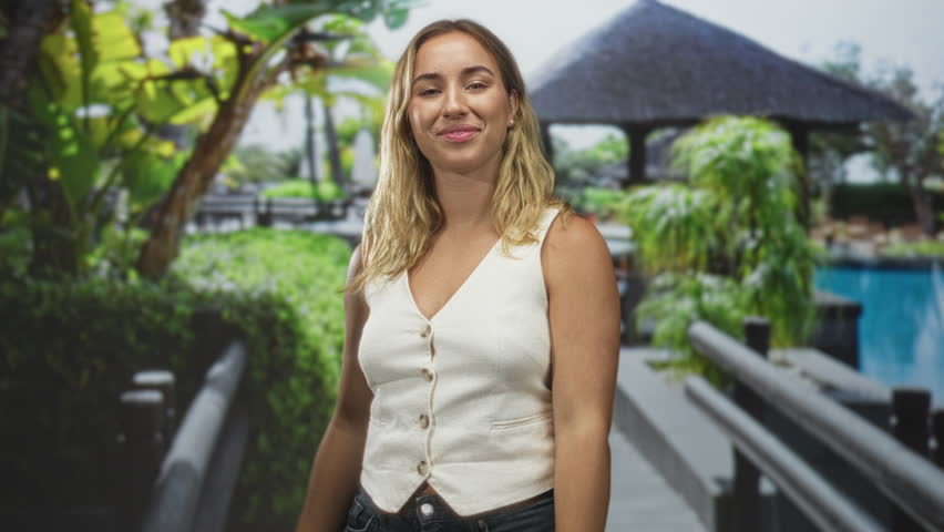 Woman pointing thumb to herself on a wooden bridge near a resort pool building, smiling and posing; playful confidence.