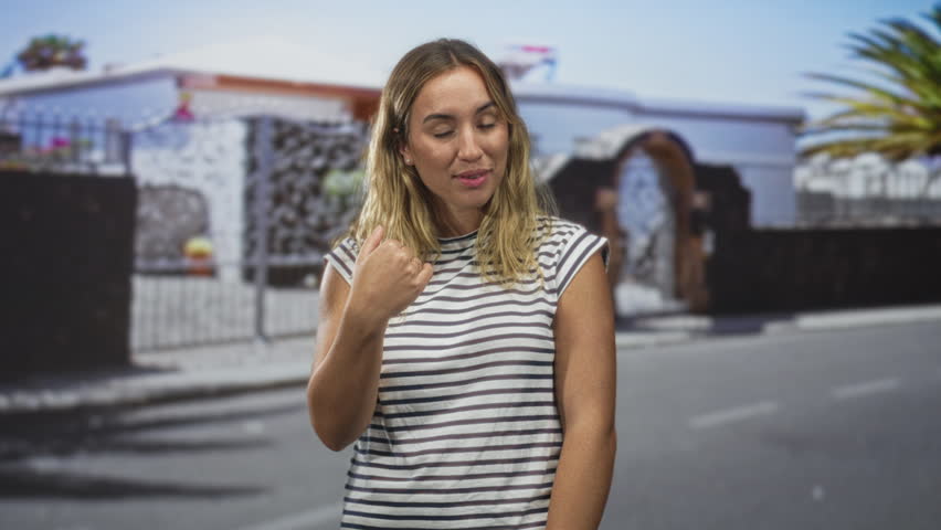 Young blonde woman smiling and showing open hands on street in front of a stone gate, wearing a striped sleeveless top; friendly invitation.