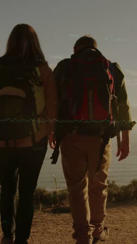 Vertical video: Two hikers hiking coast trail reaching view, shifting packs and raising arms to sea. Backlit, ocean, seascape, couple, path, backpack, vertical video