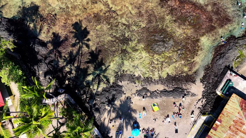 Aerial view of Kahaluu Beach Park featuring people on the sandy shore, palm tree shadows, and rocky coastline in Kailua-Kona, Hawaii, United States.