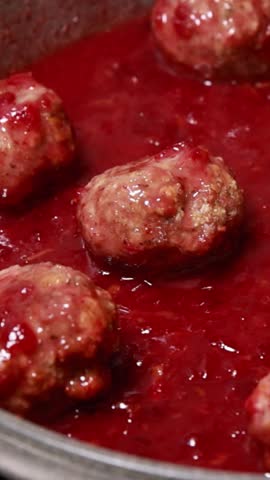 Vertical close-up of a silver spoon serving a juicy turkey meatball topped with vibrant, glistening cranberry sauce onto a white plate
