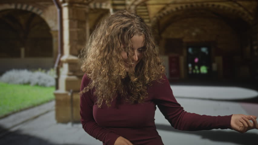 Woman with curly hair gesturing with outstretched hand and smiling in a stone arched building courtyard; playful confidence.