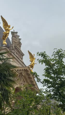 Two gilded winged allegorical figures standing atop the ornate stone facade of Petit Palais, Paris, framed by green foliage.