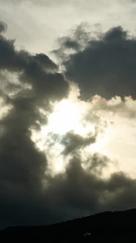 Dark, ominous storm clouds moving rapidly across the sky, obscuring the bright sun and creating a dramatic, moody atmosphere over a silhouetted hill