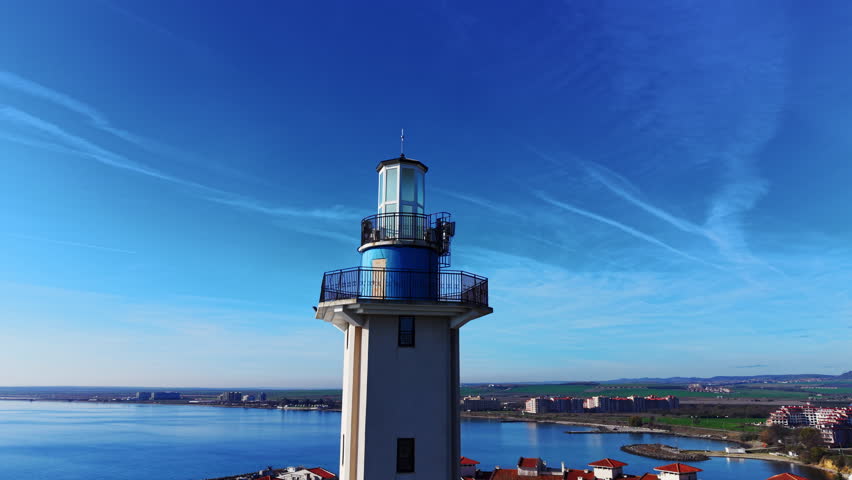 Upper part of a lighthouse featuring glass lantern room, metal railings and blue paint against bright sky. Close up of lighthouse top with blue sky background.