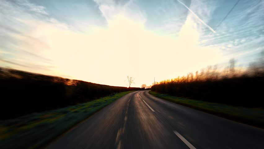 Car driving along curved countryside road at sunset through hedgerows, rural travel journey first person view. Smooth asphalt lane turning across open landscape under warm evening sky, road trip