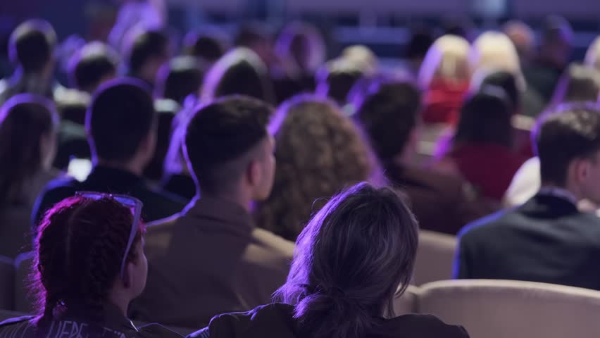 Audience seated in conference hall listening to speaker, purple stage lighting creates focused, contemplative atmosphere. Back view of diverse crowd in rows of seats.