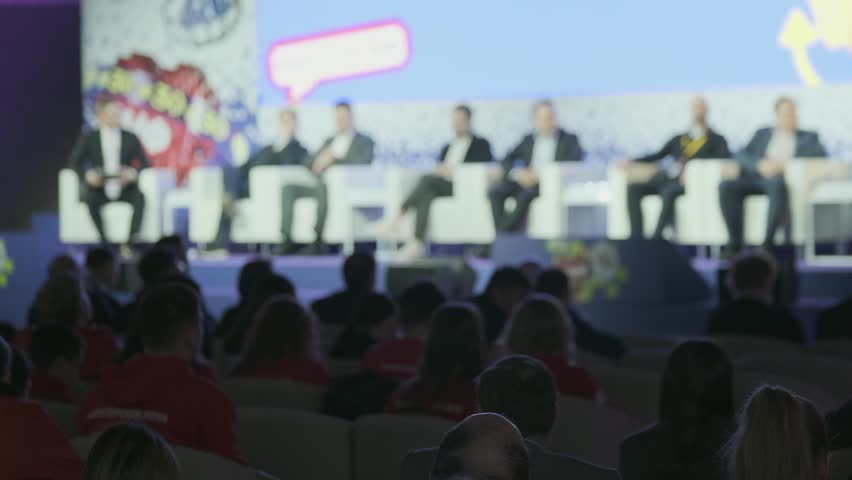 Business leaders sit on stage for panel discussion while audience members listen in dim conference hall.