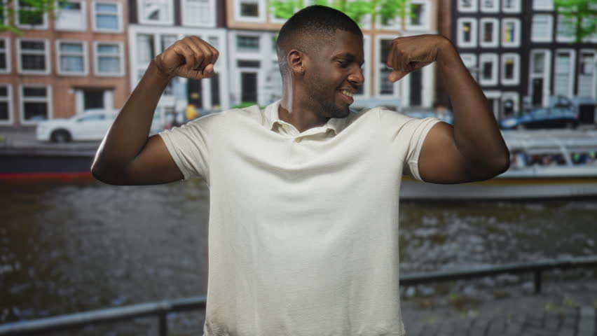 Man flexes biceps and points to arm on a street by a canal with boat and row of buildings; strength success confidence.