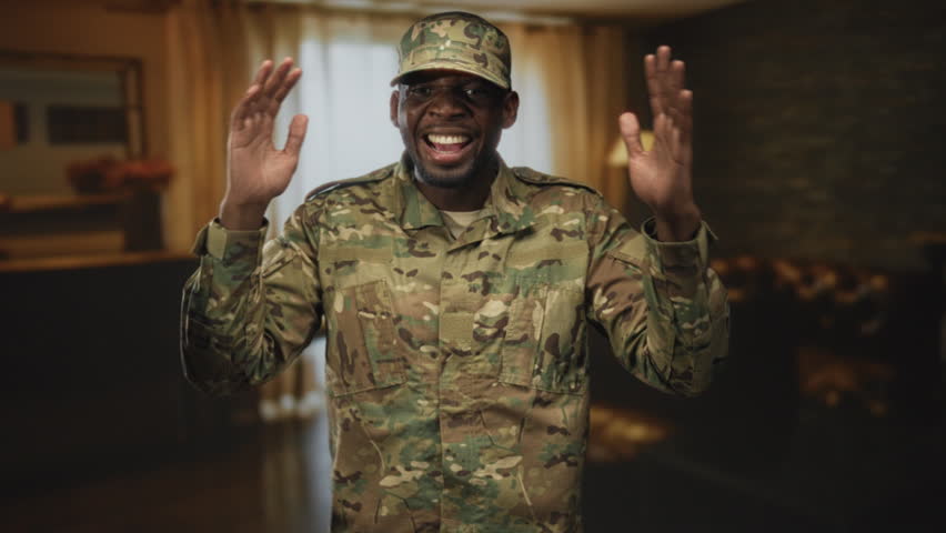Soldier in camouflage uniform raises both hands, smiling with open palms and visible teeth, cap on, staged living room set in studio; pride duty service.
