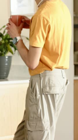 Vertical video: Glancing at art, man in orange tee talking, holding orange mug in kitchen by plant. Adult, casual, interior, minimalist, sunlit, countertop, vertical video