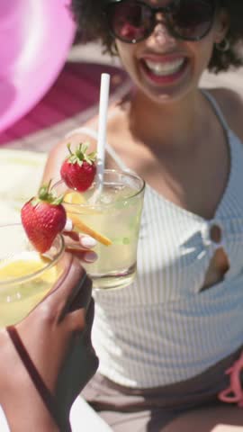 Vertical video: Holding glass to camera woman in striped top tapping straw, sipping with pal pool. Cocktail, citrus, sunglasses, deck, outdoor, summer, vertical video