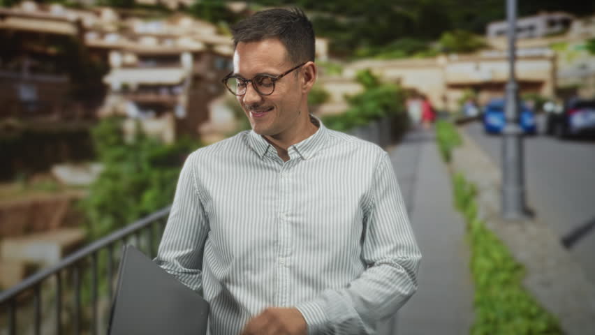 Man holding laptop with both hands adjusting screen on a street railing, typing and smiling while checking display; focus productivity.