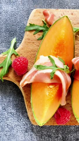 slices of fresh cantaloupe melon with italian prosciutto ham and arugula on wooden board, summer dining food