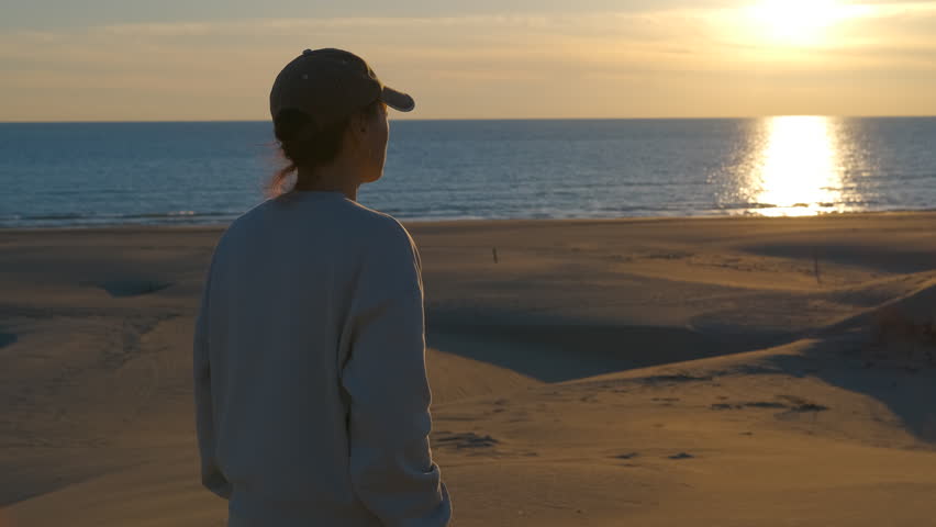 Young woman watching the sunset over sand dunes. Thoughtful young woman wearing a cap standing on a sand dune at the beach and looking at the beautiful golden sunset reflecting on the calm sea