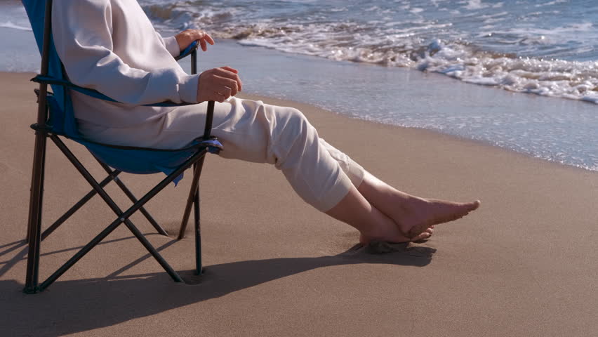 Woman relaxing on beach chair by the ocean. Young woman sitting on a folding chair relaxing on the sandy beach, with gentle ocean waves washing over her bare feet during a peaceful sunny day