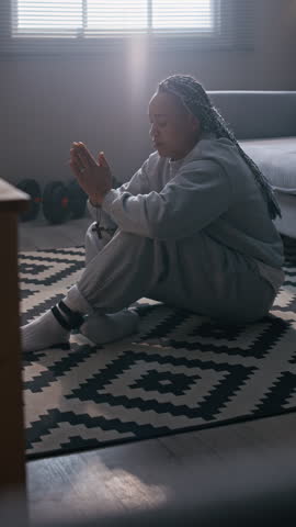 Vertical side view shot of Biracial woman with braided hair wearing electronic ankle monitor sitting on apartment floor with hands in prayer during quiet spiritual moment