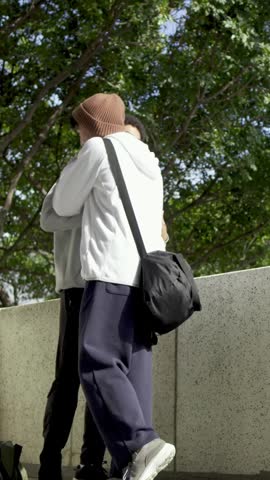 Vertical video: Passing left male in hoodie taking football after greeting by wall, beanie smiling. Outdoor, park, friends, candid, ball, handoff, vertical video