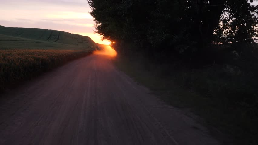 This 4K video shows a rural roadway through a countryside landscape during sunset. The visual documents silhouetted trees and the illumination of atmospheric particulates by low-angle solar light. The composition illustrates the terrestrial topography of rolling fields and transit infrastructure, documenting geographic terrain and solar transitions for editorial and creative media.
