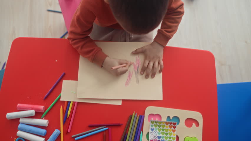 Boy drawing on paper with colored pencil, hand placed on page at a red kids table inside a building playroom; concentration creative play.