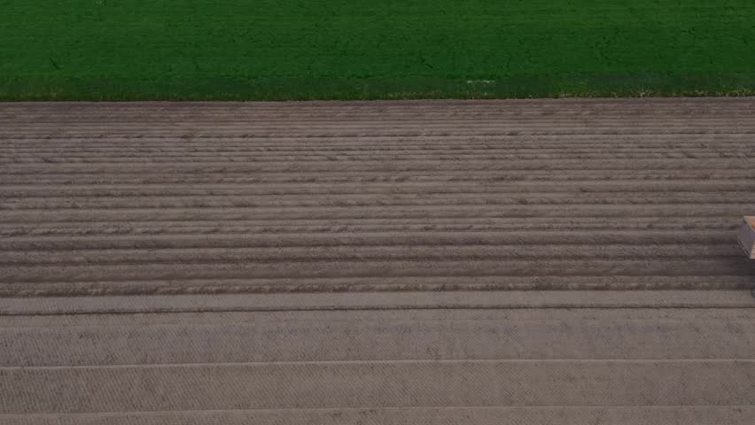 Aerial view of a blue tractor planting crops in a brown furrowed field next to a green meadow in Middenbeemster, North Holland, Netherlands.