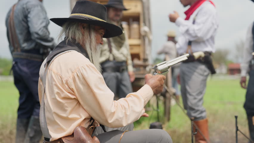 An old man sit showing off, brandishing a gun, in front of a cowboy caravan with day light.