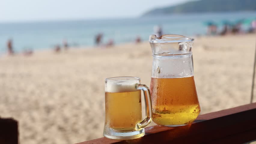 Cold beer pitcher and mug beachside, ocean view, people relaxing.
