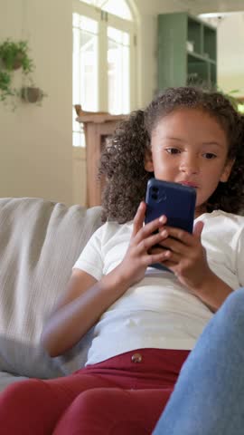 Vertical video: Tapping girl white tee at home with blue phone reacting as adult shows other phone. Child, family, smartphone, couch, livingroom, candid, vertical video