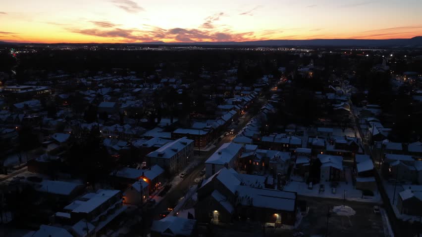 Snow covered residential neighborhood in Pennsylvania at sunset with glowing street lights and winter atmosphere. Calm suburban evening mood with cold seasonal landscape. Aerial rise wide shot.