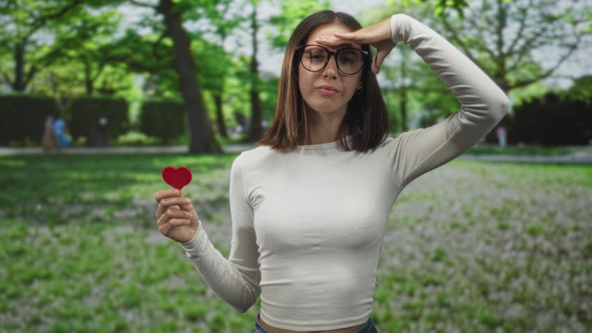 Young hispanic woman holds small red felt heart prop at chest, hand shading eyes while smiling gently in forest; joy love.