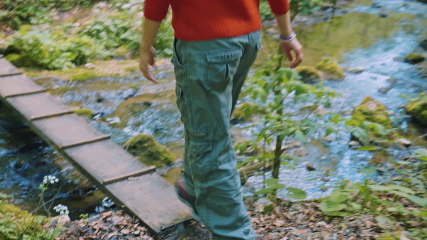 A young woman with white hair tied back in a ponytail walks through a lush green forest, her red hoodie contrasting against the vibrant spring foliage.