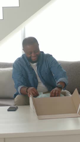 Vertical video: Opening box, man in denim checking black shoes on sofa, light-blue tissue showing. Footwear, package, candid, backlit, natural, minimalist, vertical video