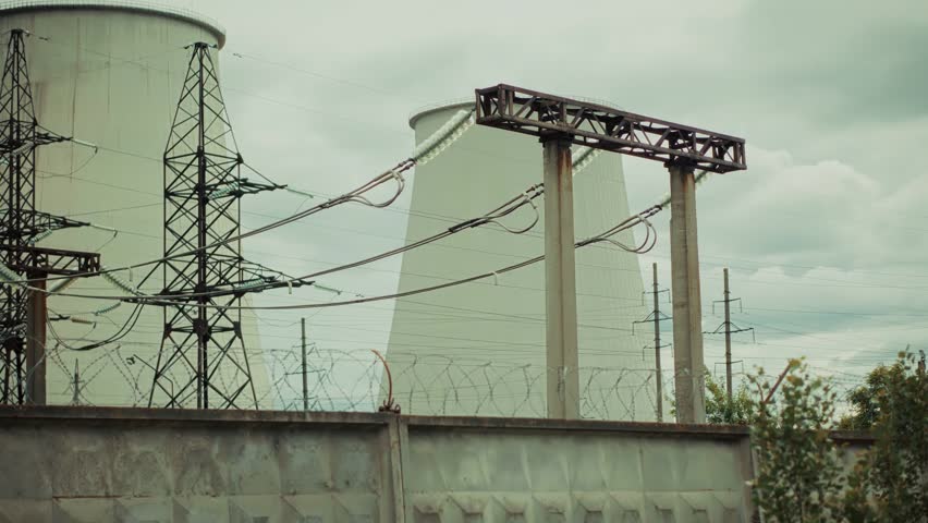 Industrial plant with large cooling towers and high voltage power lines behind a fence. Electrical infrastructure provides energy for urban development during a cloudy day. Every station requires regular technical maintenance for stable facility performance. This power environment ensures reliability for global electricity production. Metal towers stand near concrete wall while wires transmit energy across the territory. Industrial zone provides background for engineering and utility services. Complex systems help achieve effective management and planning for the city. Massive construction supports energy distribution