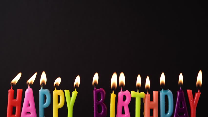 Birthday candles glow brightly against black background. Colorful letters spell Happy Birthday. Scene radiates birthday celebration.