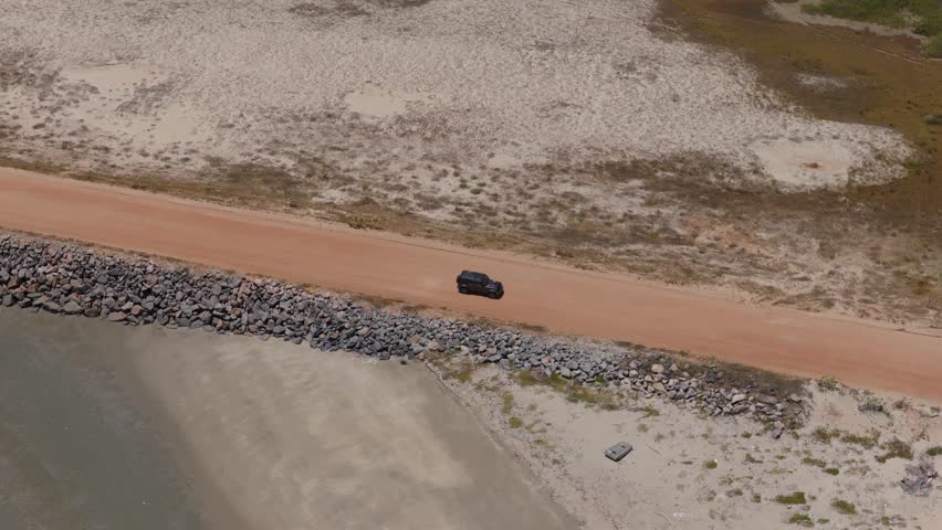 A professional aerial panning shot captures a dark SUV traveling along a red-clay road near Ilha do Guajiru, Brazil. A towering white wind turbine dominates the foreground against the dunes.