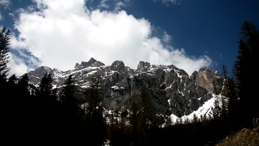 Dramatic clouds move over rocky alpine peaks with dark pine forest foreground