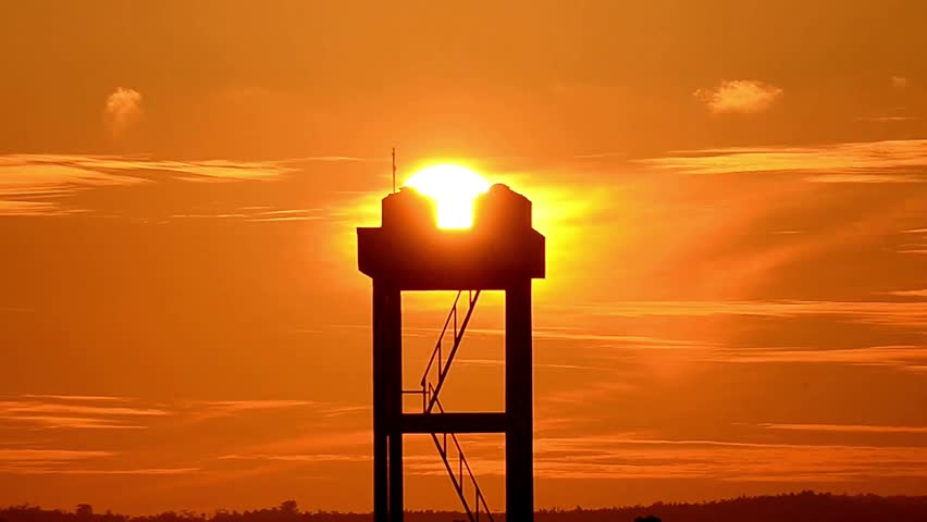 The setting sun aligns with a tall silhouetted tower amid glowing orange clouds