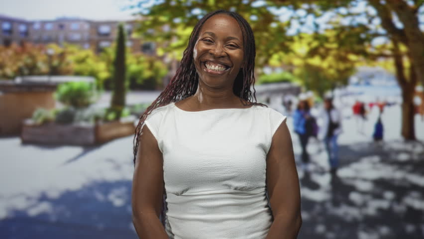 African american woman in a white dress framing her face with fingers and smiling on a sunny street; joyful playfulness.