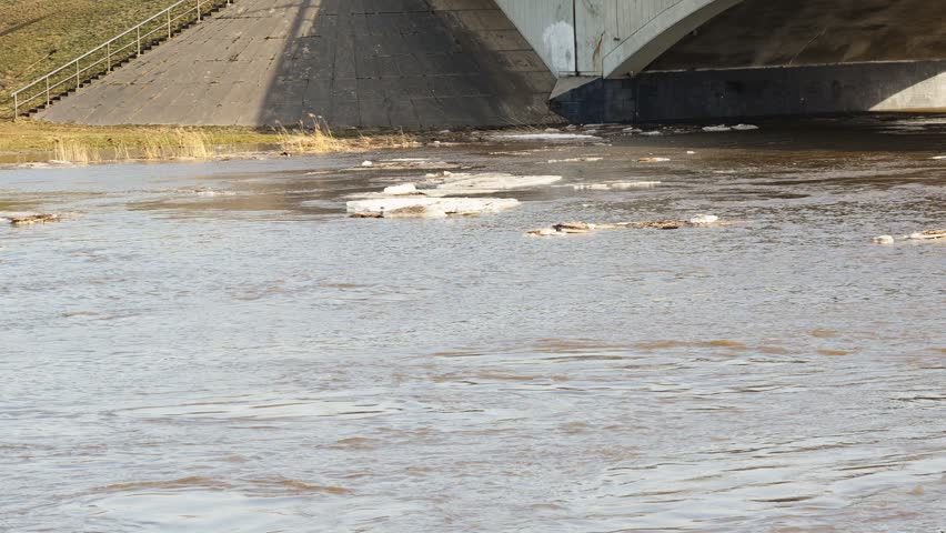 Melting ice floes drift slowly along the current of a wide, muddy river. A large concrete bridge structure and grassy riverbank are visible in the background