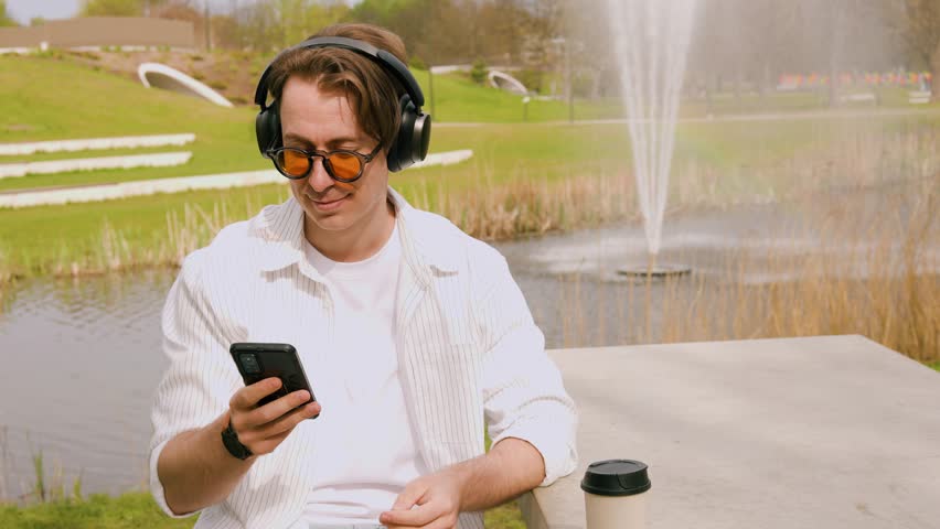 A young man in orange sunglasses enjoys music through headphones in a city park. 4K video featuring a sunny day, park fountain, and outdoor relaxation. High quality footage