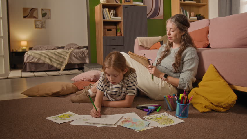 Low eye level shot of preteen girl lying on floor drawing pictures with colorful crayons while chatting with mother during shared activity in living room
