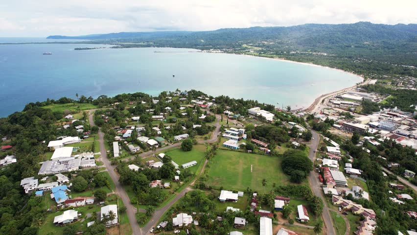 Aerial view of the coastal town of Wewak featuring residential houses, lush green hills, and a large bay under a cloudy sky in Wewak, East Sepik Province, Papua New Guinea.