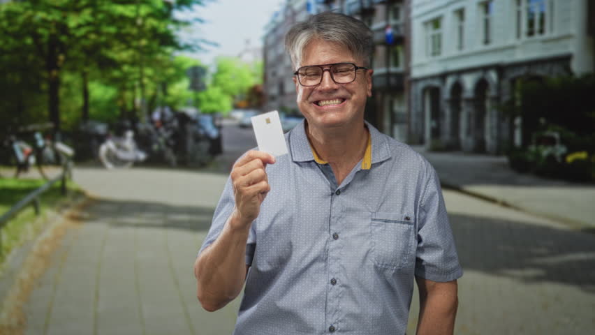 Man with grey hair holding a plain creditcard, smiling and pointing finger toward the right on a tree lined street; trust confidence promotion.