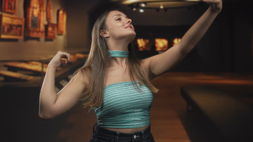 Young woman taking selfie with smartphone, arm extended and puckering lips in museum building; playful memory.