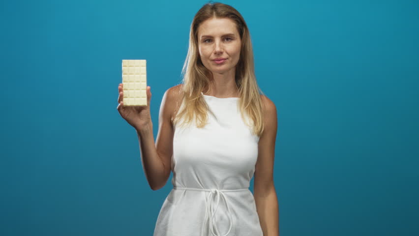 Blonde woman holding white chocolate bar, points finger toward camera while smiling and wearing white dress in studio; playful temptation indulgence.