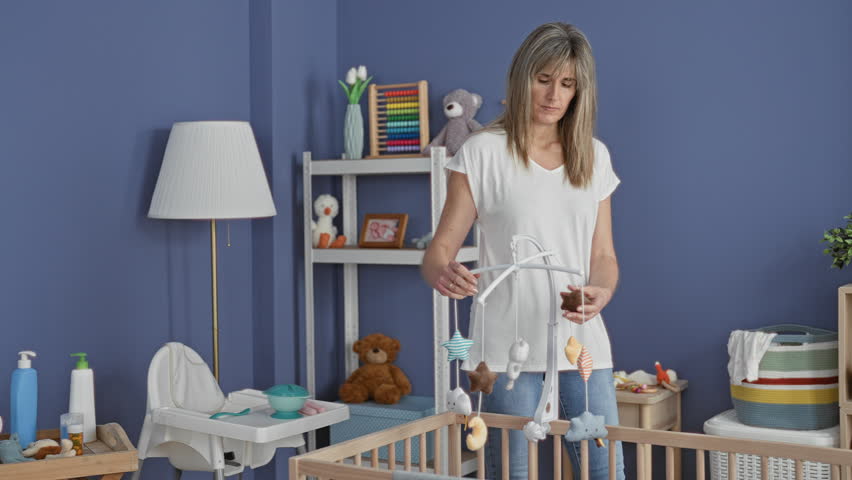 Woman holding a baby mobile by hand above a wooden cradle in a nursery, adjusting plush stars and moons while gazing down; serenity.